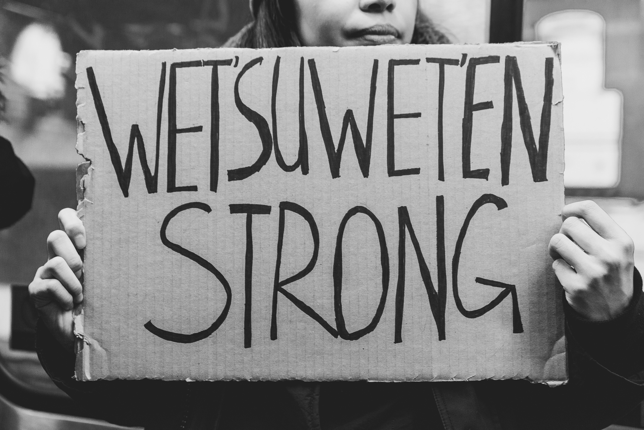 A woman holding a cardboard sign with 'Wetsuweten Strong' written on it in big letters.