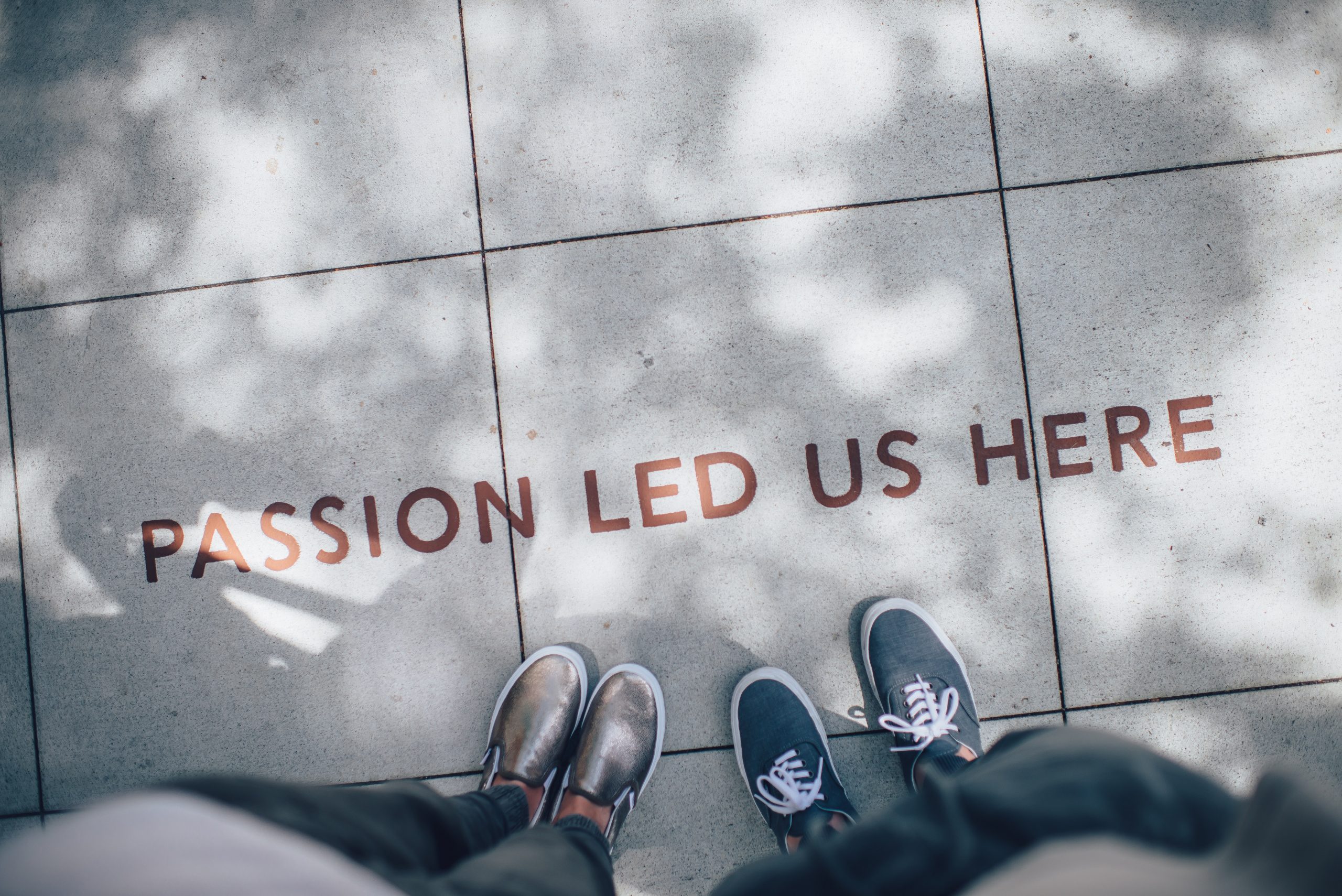 A photo looking down at two people standing over a quote painted on the sidewalk that reads "Passion Led Us Here"