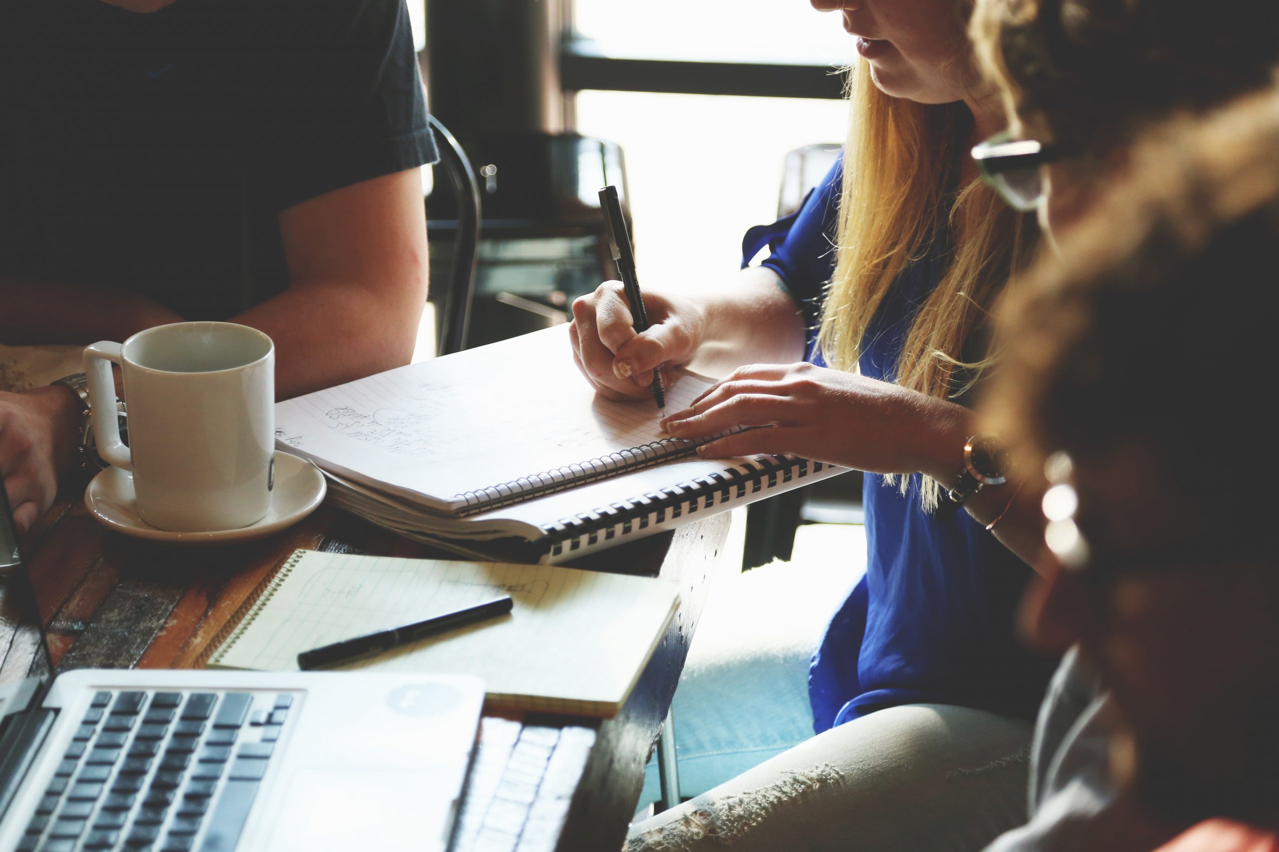 Image of four people gathered at a table, working on a laptop and writing on notebooks and notepads