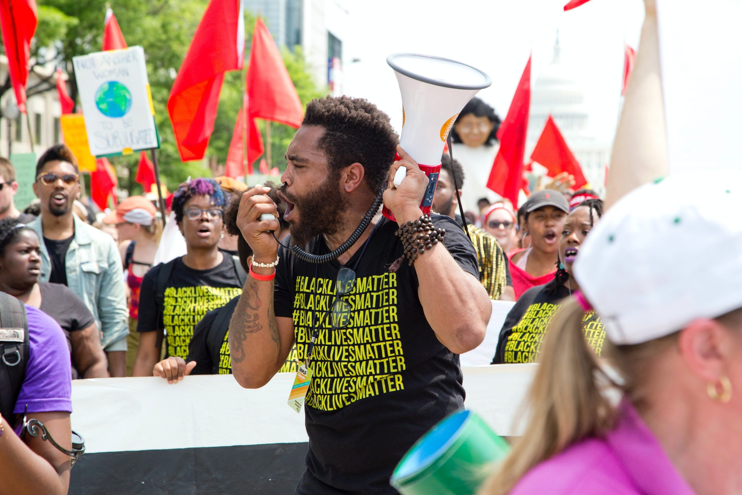 A Black man wearing a Black Lives Matter T-shirt speaks into a megaphone to a crowd.