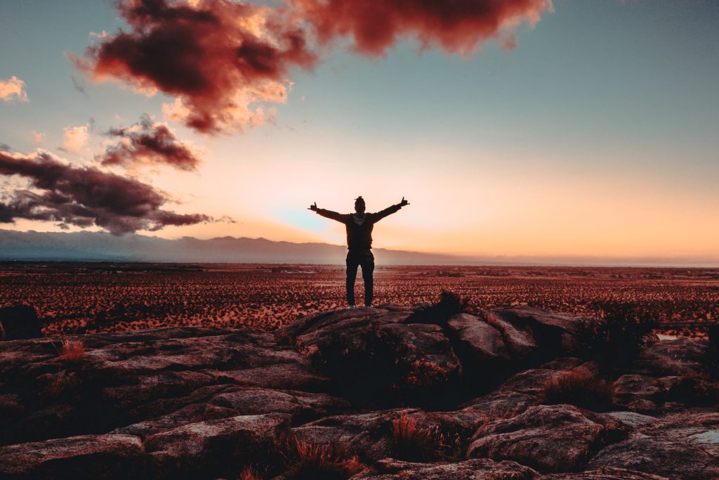A person standing on a hill overlooking a vast landscape at sunset with their hands open wide as a sign of achievement.