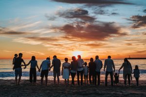 Photograph of a large group of people standing side by side on a beach watching the sun set into the ocean. People are holding hands and have their arms around one another.