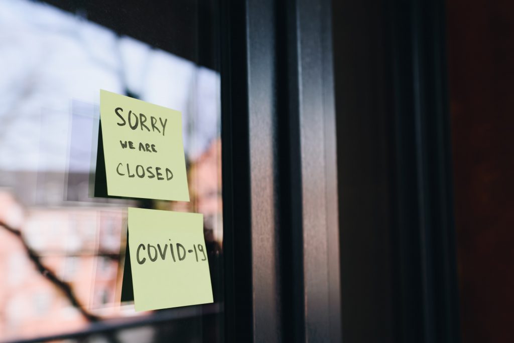 Image of a retail store's front door with a post-it-note on the front reading, "sorry we are closed....covid-19".