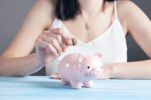 Image of a woman putting a coin into a traditional-looking piggy bank (small pink pig that holds money).