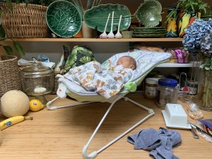 "A picture of a baby in a baby chair on top of a kitchen counter. Garlic, a banana, a melon, green dishes, and baskets are also on the counter."