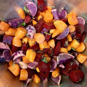 "A metal bowl shown directly from above with chopped beets, carrots, yams, and herbs inside."