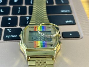 "Image of a computer keyboard. A Timex watch is resting on the keys. The watch has a rainbow pattern. On top of the watch is a ring."