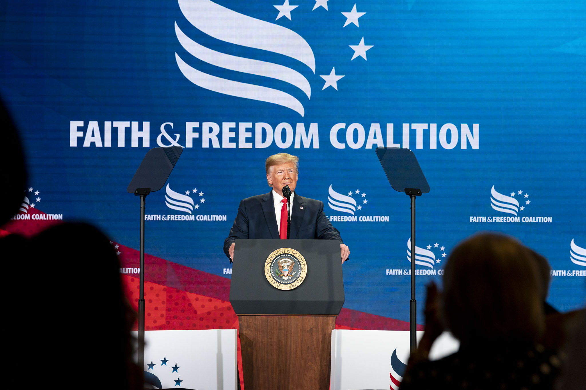 Smiling man in suit and red tie stands behind a podium with the American Seal. Behind him a blue poster reads faith and freedom coalition