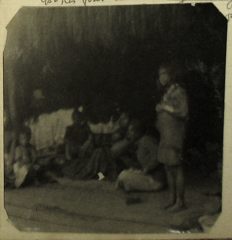 Blurry photograph, several seated figures in the background, one standing figure in the foreground.