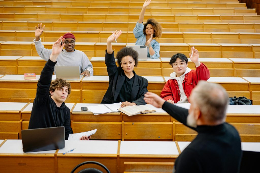 Students of diverse backgrounds raising their hands in a classroom, actively participating in response to a teacher's question, reflecting an inclusive and respectful learning environment.