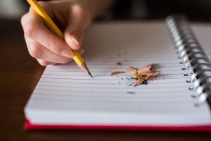 A person writes in a notebook with a pencil. Pencil shavings are on the notebook page.